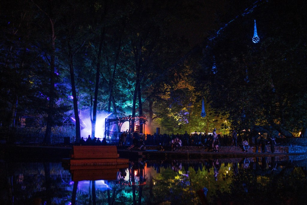 Orbits festival in the forest by the river. Colored lights and lasers permeate the haze and reflect on the river waters. People hang by the beach.