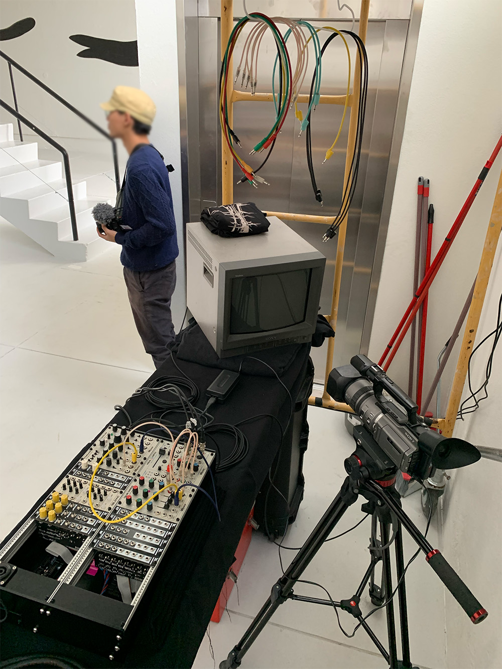 Video gear. On top of a table, a modular video system with cable spaghetti connecting modules and a cathode ray tube television. A camera stands on a tripod pointing towards the tv. The space is luminous, very white. The gear is mostly dark colored — grey and black.